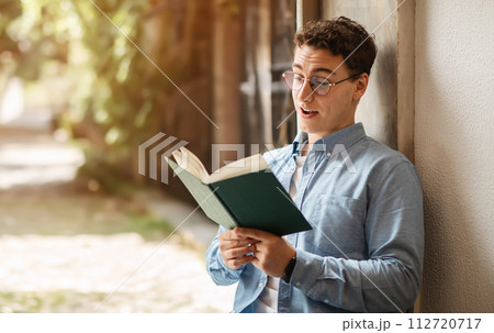 Interested young man with curly hair and glasses engrossed in reading a book 112720717