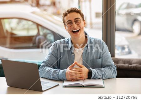 Laughing young man with curly hair and glasses sitting with hands clasped in front of his open notebook Laughing young man with curly hair and glasses sitting with hands clasped in front of his open notebook 112720768