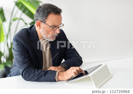 Serious and focused mature businessman with glasses intently using a tablet Serious and focused mature businessman with glasses intently using a tablet 112720868