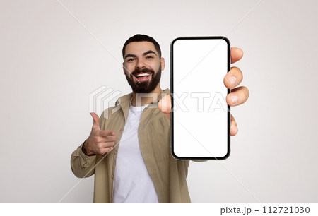 Enthusiastic young man with a beard, in a khaki shirt, cheerfully holds a smartphone with a blank screen Enthusiastic young man with a beard, in a khaki shirt, cheerfully holds a smartphone with a blank screen 112721030
