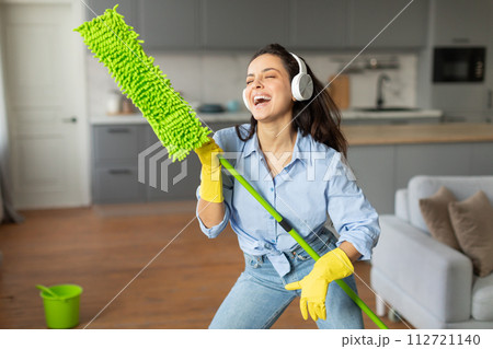 Happy woman with headphones using mop playfully Happy woman with headphones using mop playfully 112721140