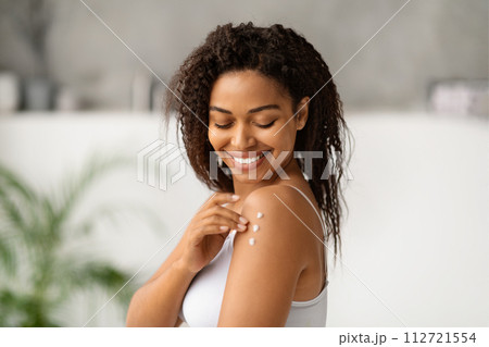 Joyful black woman smiling while applying body lotion to her shoulder 112721554