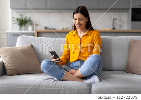 Smiling young woman browsing on phone while sitting on sofa 112722079