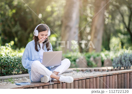 A content young woman enjoys learning on her laptop outdoors, surrounded by nature 112722120