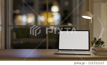 A wooden desk features a laptop, books, a decor plant, and a table lamp in a cosy room at night. 112722127