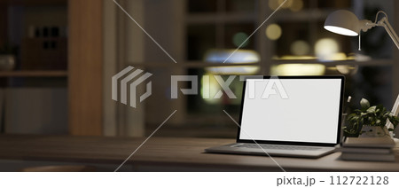 A wooden desk features a laptop, books, a decor plant, and a table lamp in a cosy room at night. 112722128