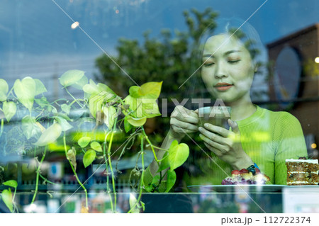 Asian woman enjoying her coffee in a cafe, viewed through a window with reflections of foliage Asian woman enjoying her coffee in a cafe, viewed through a window with reflections of foliage 112722374
