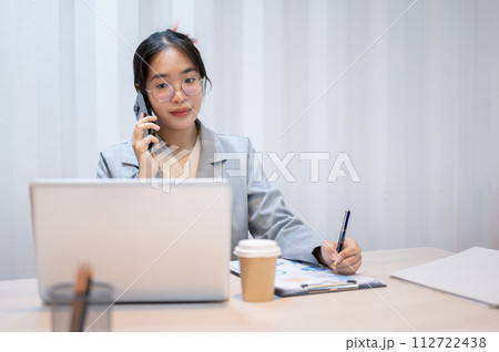 A businesswoman is talking on the phone, taking notes, and reading information on her laptop. A businesswoman is talking on the phone, taking notes, and reading information on her laptop. 112722438