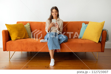 Content young woman in casual wear smiling while using smartphone at home Content young woman in casual wear smiling while using smartphone at home 112723007
