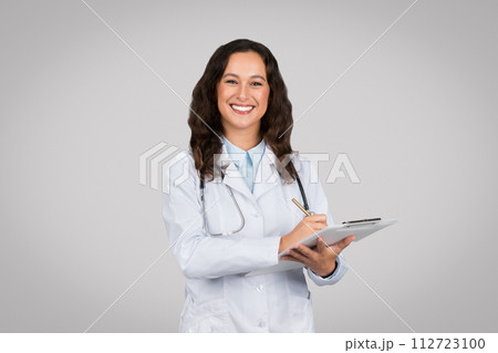 Cheerful woman doctor in white lab coat writing on clipboard with pen, grey studio background Cheerful woman doctor in white lab coat writing on clipboard with pen, grey studio background 112723100