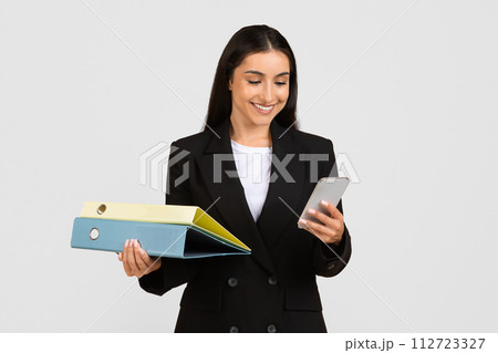 Smiling businesswoman multitasking with folders and smartphone Smiling businesswoman multitasking with folders and smartphone 112723327