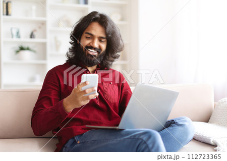 Cheerful indian guy sitting on couch at home, using gadgets Cheerful indian guy sitting on couch at home, using gadgets 112723365