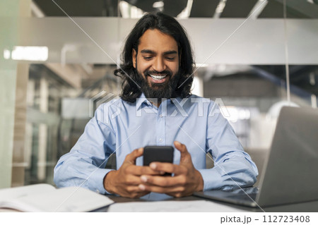 Positive indian guy manager using smartphone, modern office interior 112723408