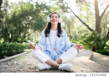 A serene woman enjoys meditation in a sunlit park, headphones on and smartphone in hand 112723409