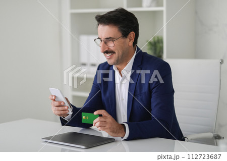 Joyous businessman in a blue suit engaging with his mobile phone while holding a debit card Joyous businessman in a blue suit engaging with his mobile phone while holding a debit card 112723687