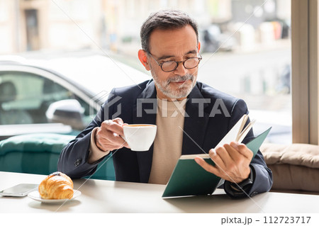 Intelligent mature man with glasses enjoying a cup of coffee while reading a book 112723717