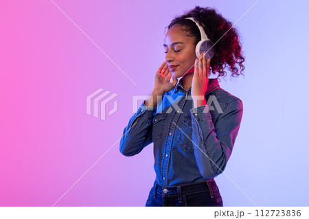 Relaxed black lady enjoying music in headphones, denim attire, colorful backdrop Relaxed black lady enjoying music in headphones, denim attire, colorful backdrop 112723836