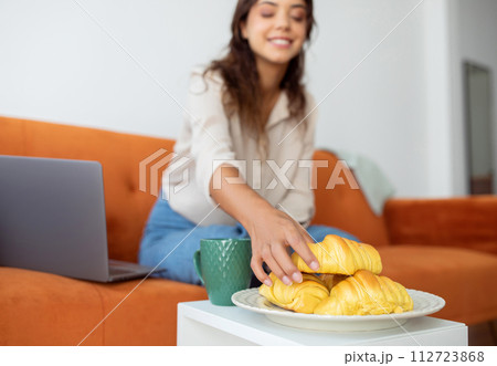 Smiling woman reaching for croissants on plate next to her laptop Smiling woman reaching for croissants on plate next to her laptop 112723868