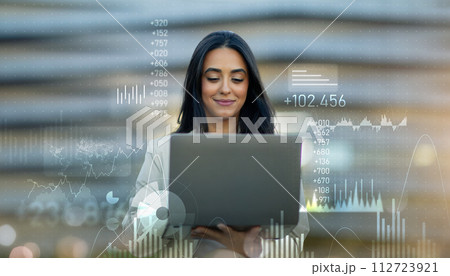 Confident young professional woman using a laptop, overlayed with dynamic financial charts 112723921