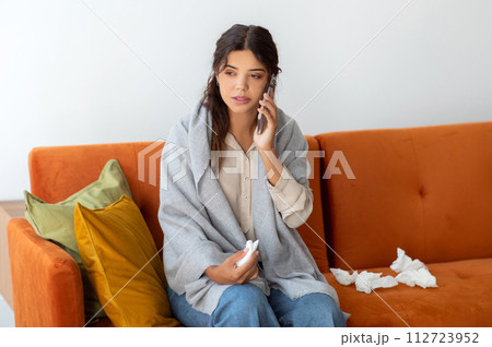 Concerned young woman wrapped in blanket making phone call to doctor Concerned young woman wrapped in blanket making phone call to doctor 112723952
