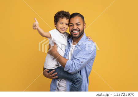 Smiling black boy showing thumbs up at camera while father holding him Smiling black boy showing thumbs up at camera while father holding him 112723970