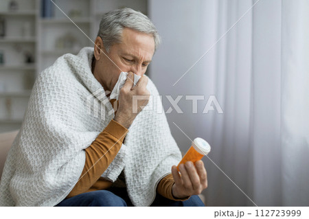 Sick elderly man sneezing and holding jar with pills 112723999