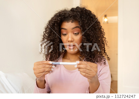 Shocked black woman checking pregnancy test at home, feeling surprised with result 112724247