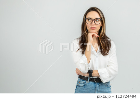 A contemplative young woman with long hair and glasses rests her chin on her hand 112724494