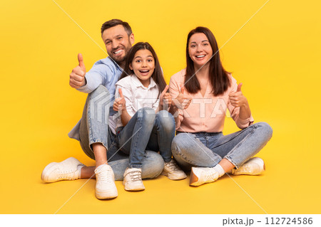 Happy family of three giving thumbs up sitting, yellow backdrop 112724586