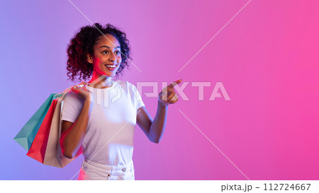 Joyful lady shopper with bags pointing to side at free space on vibrant pink and blue background 112724667