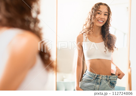 Caucasian teenager girl at mirror showcasing weight loss success indoors Caucasian teenager girl at mirror showcasing weight loss success indoors 112724806