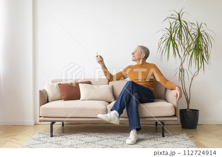 Senior man sitting on couch at home, turn on AC Senior man sitting on couch at home, turn on AC 112724914