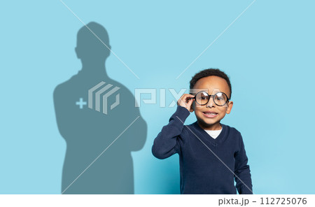 A confident young boy with glasses adjusts his frames, smiling against a blue background A confident young boy with glasses adjusts his frames, smiling against a blue background 112725076