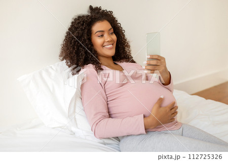 Smiling black pregnant woman comfortably reclining in bed and using smartphone Smiling black pregnant woman comfortably reclining in bed and using smartphone 112725326