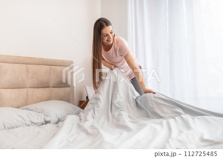 Cheerful woman making bed in bright room, displaying everyday domestic life Cheerful woman making bed in bright room, displaying everyday domestic life 112725485