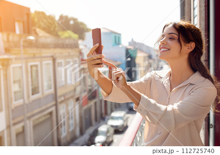 Smiling Young Woman Standing On Balcony And Taking Photo Of Cityscape 112725740