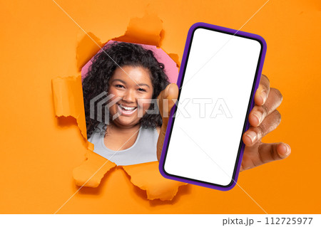 Cheerful young woman with curly hair peering through a torn orange paper, presenting a smartphone with a blank screen Cheerful young woman with curly hair peering through a torn orange paper, presenting a smartphone with a blank screen 112725977