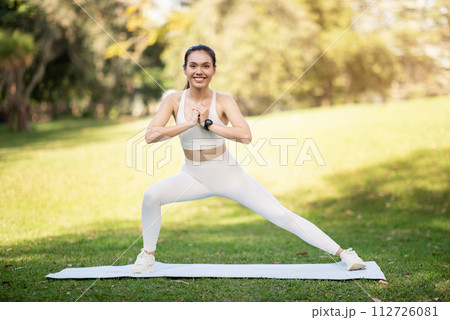 A radiant young woman in a white sports outfit performs a dynamic lunge on a yoga mat 112726081