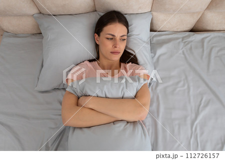Upset young woman lying in bed, hugging pillow with a worried expression Upset young woman lying in bed, hugging pillow with a worried expression 112726157