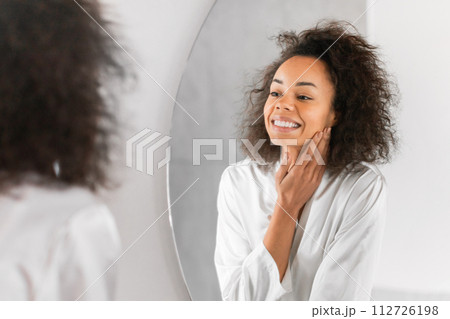 Smiling black woman looks at mirror enjoying flawless skin indoors Smiling black woman looks at mirror enjoying flawless skin indoors 112726198