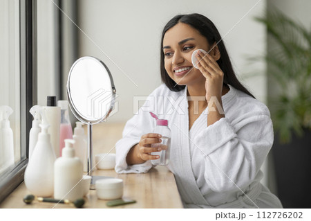Positive young indian woman cleansing face in bathroom Positive young indian woman cleansing face in bathroom 112726202
