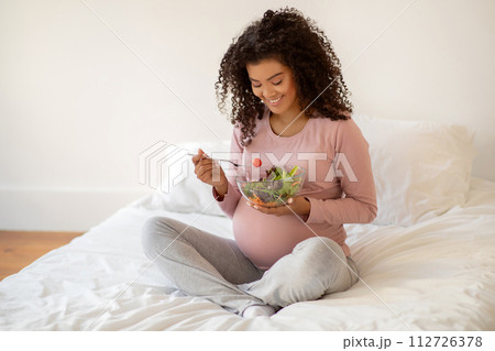 Beautiful black pregnant woman sitting on bed and holding bowl of salad 112726378
