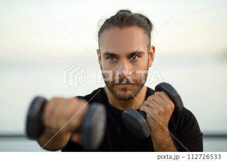 Determined man in fitwear exercises with dumbbells flexing biceps outside 112726533