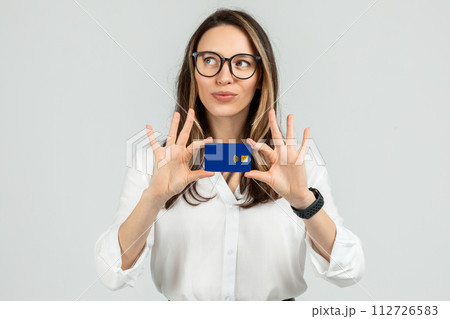 Curious young woman with glasses examining a blue credit card, wearing a white blouse 112726583
