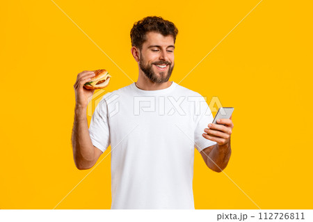 Cheerful young man holding burger using cellphone over yellow background 112726811