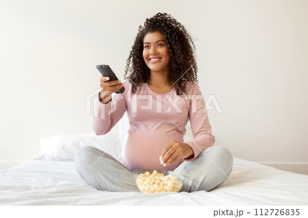Pregnant black woman in pink shirt enjoying popcorn and holding remote controller 112726835