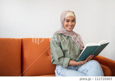 muslim lady in headscarf sits on sofa with book indoors 112726865
