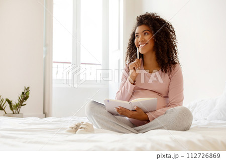 Happy Black Pregnant Woman Writing Checklist In Notepad While Sitting On Bed Happy Black Pregnant Woman Writing Checklist In Notepad While Sitting On Bed 112726869