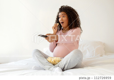 Shocked pregnant woman with popcorn and remote reacting to content on tv 112726959