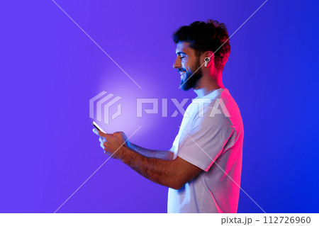 Young caucasian man holds phone enjoying music through earbuds, studio 112726960